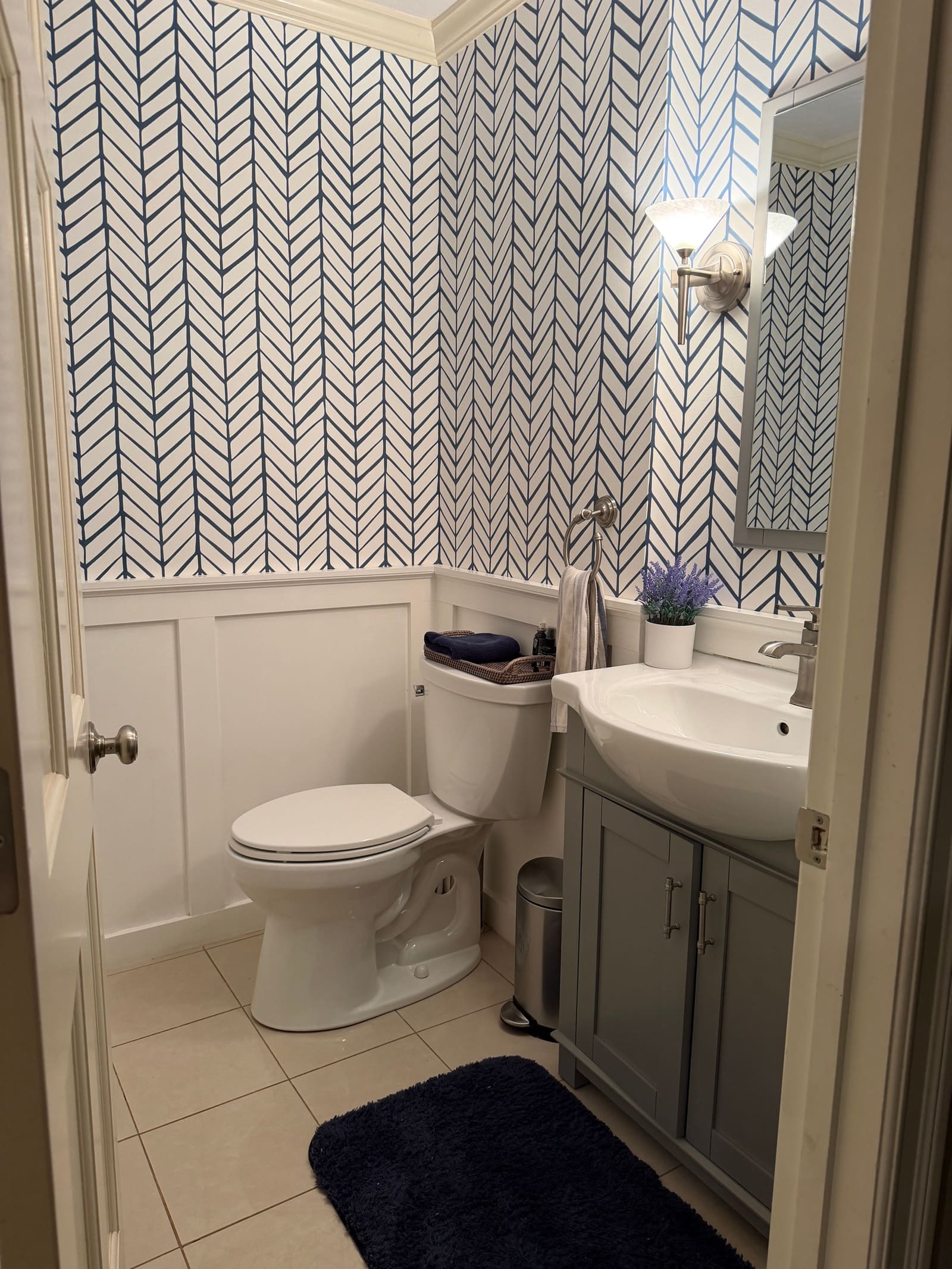 A finished powder room with herringbone wallpaper over wainscoting and a gray vanity